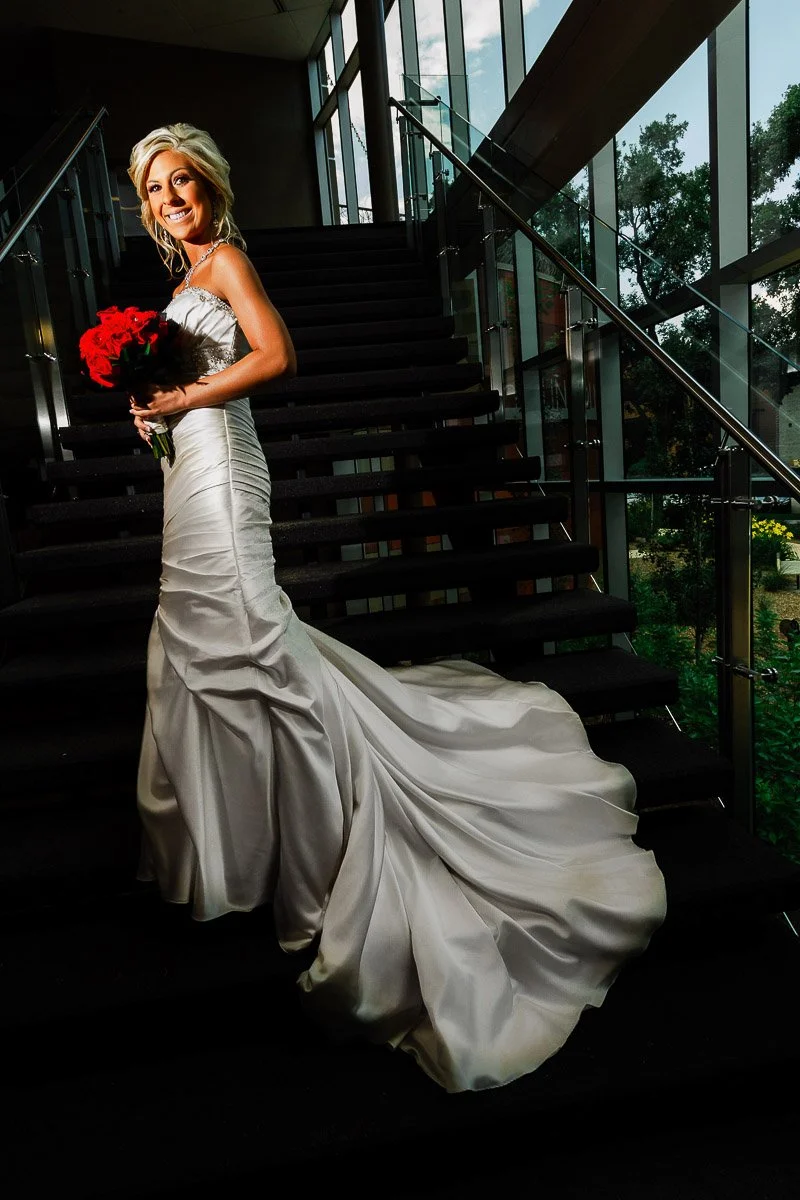 A bride in a satin gown with a long train holds a bouquet of red roses, standing on a grand staircase during a Lincoln Center wedding with glass railings and large windows.