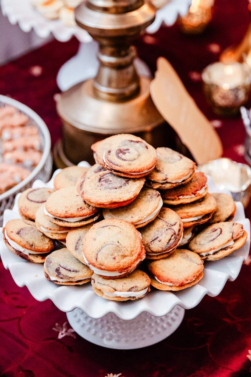 A tower of swirled cookies with cream filling on a decorative white stand, set against an elegant table with warm lighting and brass accents.