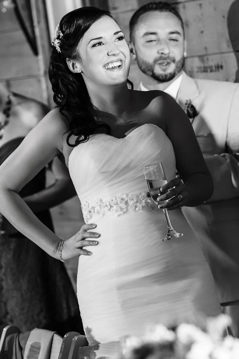 A bride in a strapless gown smiles joyfully, holding a champagne glass, radiating happiness. A man behind her looks on warmly. The scene conveys celebration.