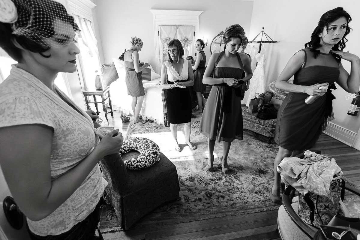 Bridesmaids in a warmly lit room prepare for a wedding, adjusting dresses and hairstyles. The scene is lively and anticipatory, with a wedding gown visible.