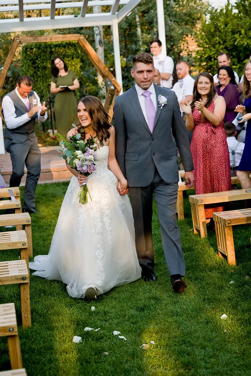 A joyful bride in a white gown and groom in a gray suit walk hand in hand down an outdoor aisle. Guests clap and smile, creating a celebratory atmosphere.