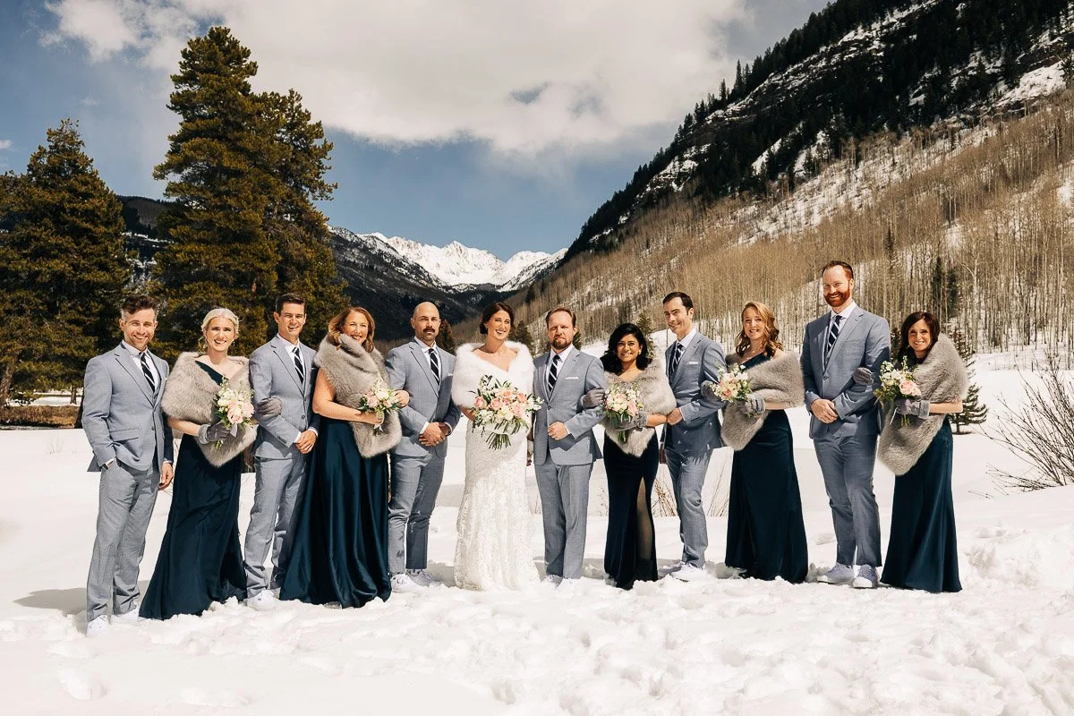 A wedding party poses smiling in a snowy mountain landscape. The bride in a white gown is surrounded by attendants in gray suits and dark dresses, creating a joyful, elegant scene.
