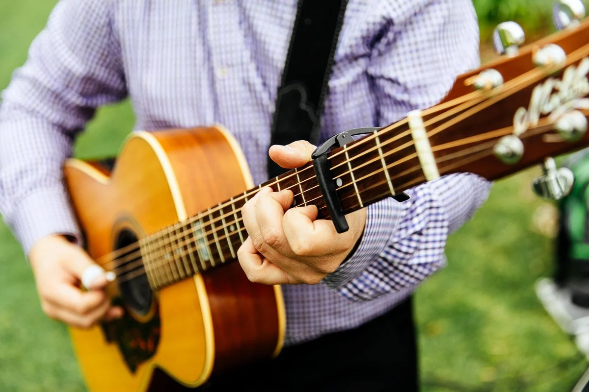 A person in a checkered shirt strums an acoustic guitar with a capo on the neck, conveying a lively, musical atmosphere in an outdoor setting.