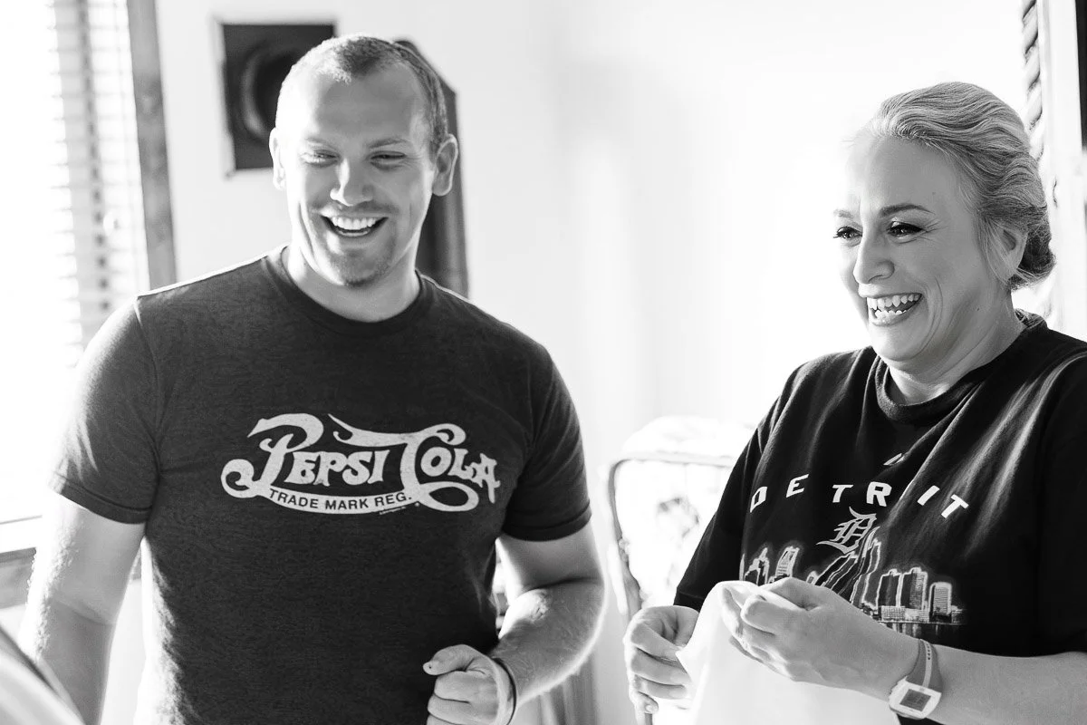 A black-and-white image of a man and woman laughing together indoors. The man wears a Pepsi Cola shirt, and the woman wears a Detroit shirt. They appear joyful and relaxed.