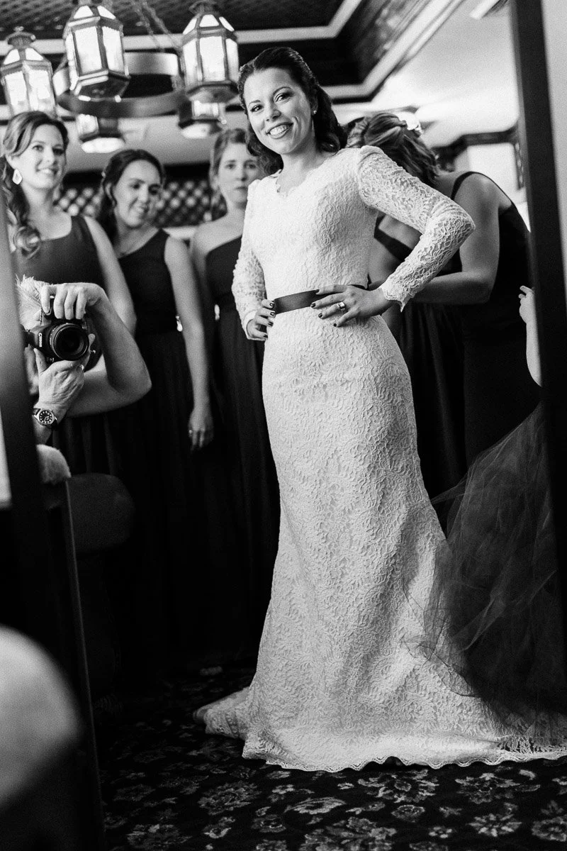 A joyful bride in an elegant lace gown poses for a photo, surrounded by bridesmaids in dark dresses. The mood is warm and celebratory.