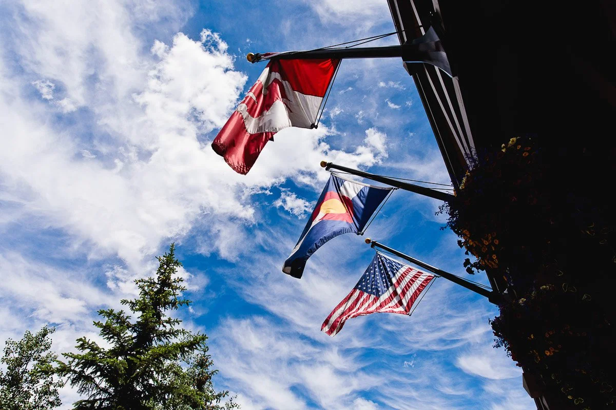 Three flags, including Canadian and American, and a state flag are waving against a blue sky with scattered clouds. A pine tree is visible below.