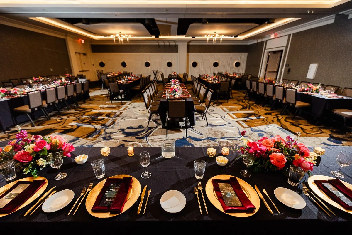 Elegant banquet hall with black tablecloths and floral centerpieces. Place settings with gold plates and red napkins. Warm, inviting atmosphere.