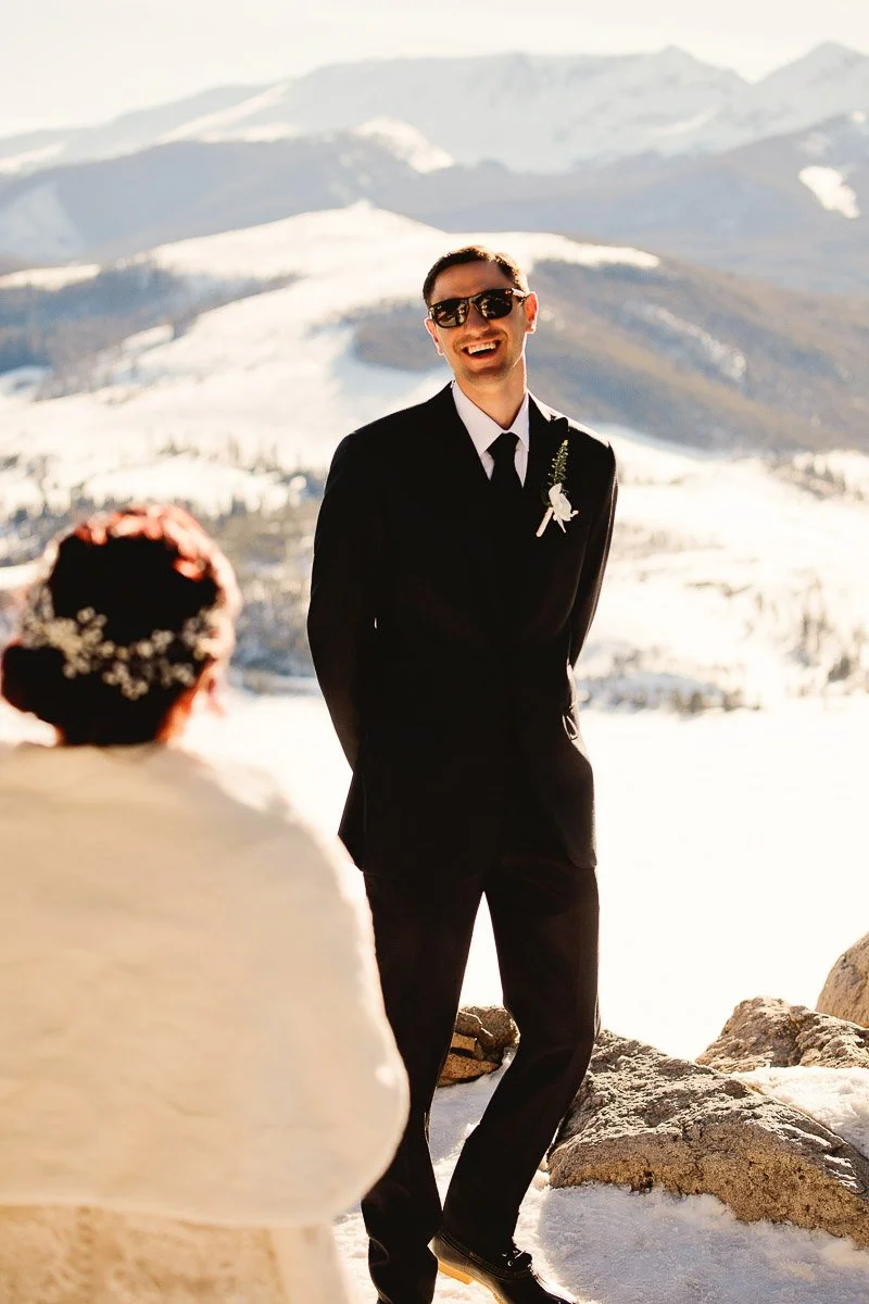 A groom in a black suit and sunglasses smiles joyfully at his bride against a snowy mountain backdrop. The scene conveys warmth and happiness.