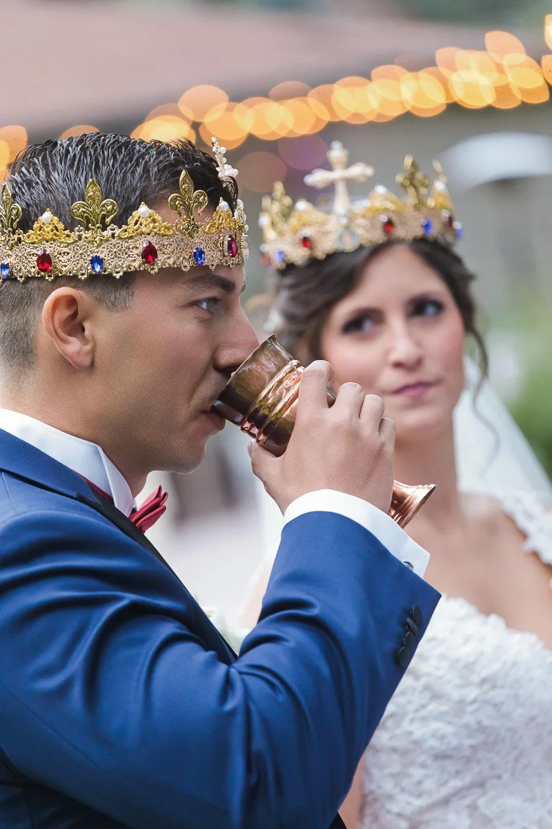 A couple wearing ornate gold crowns decorated with jewels participates in a wedding ceremony. The groom in a blue suit drinks from a chalice, while the bride in a white dress looks on. Festive lights are blurred in the background, adding a celebrator
