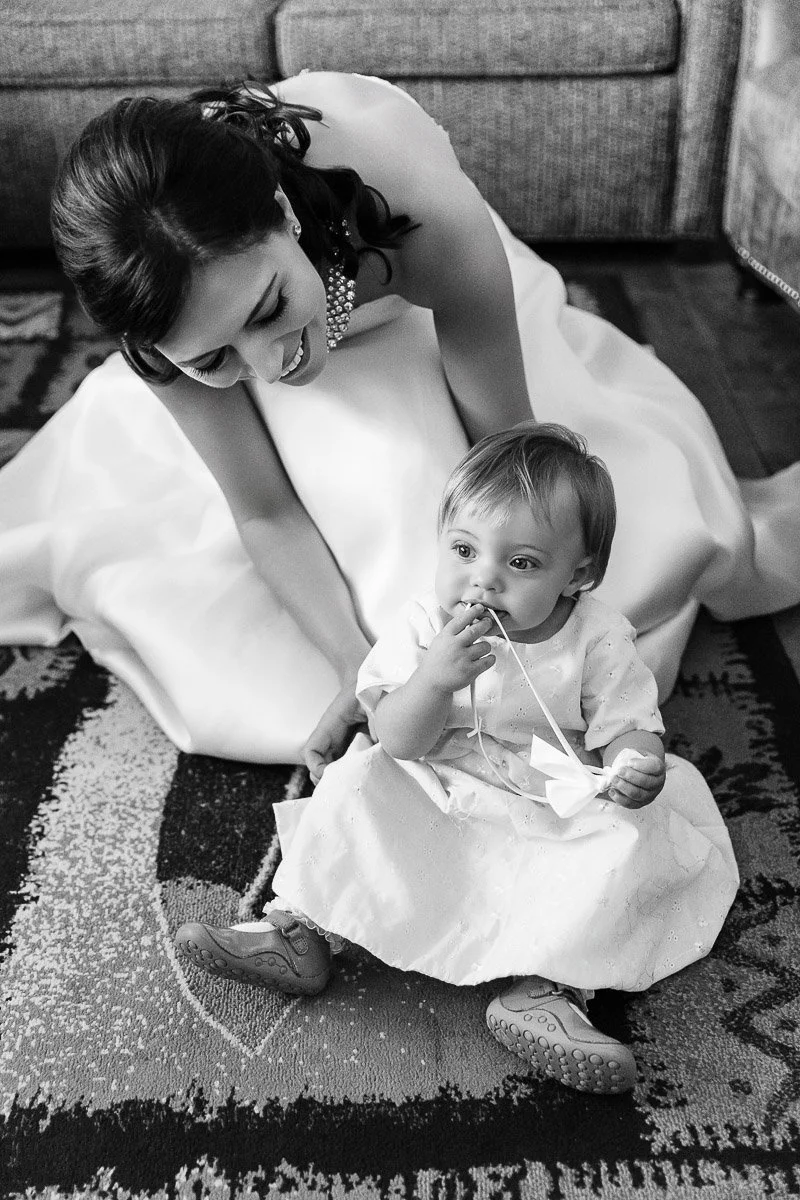 A woman in a white dress kneels on a patterned rug, smiling affectionately at a baby sitting in front of her, who is holding a ribbon. The scene conveys warmth and joy.