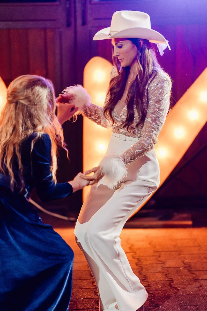 Two women joyfully dance at an event. One wears a cowboy hat and chic white outfit, while the other wears a blue dress. Warm lights glow behind them.