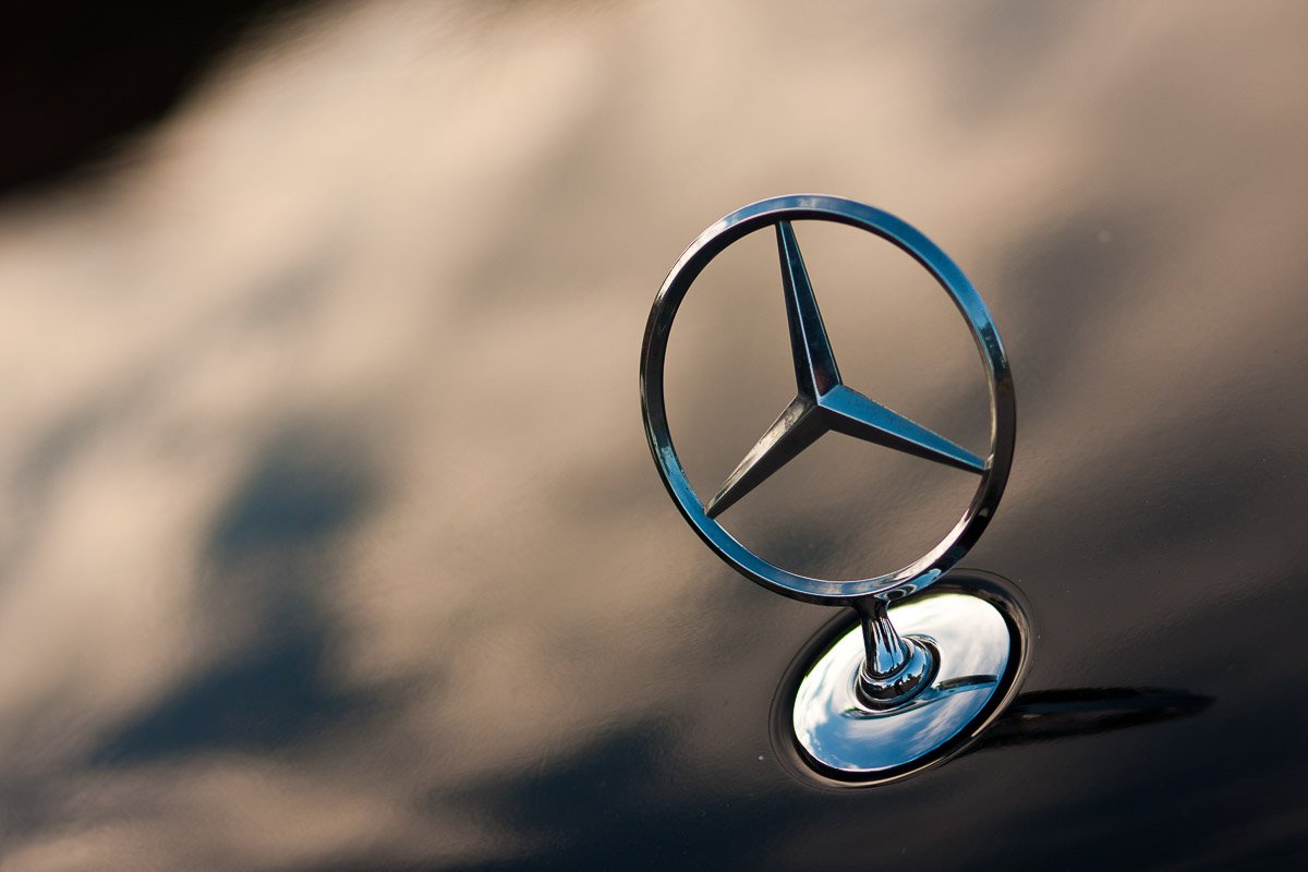 Close-up of a Mercedes-Benz emblem on a car hood, set against a soft sky reflecting on the shiny black surface, conveying elegance and luxury.