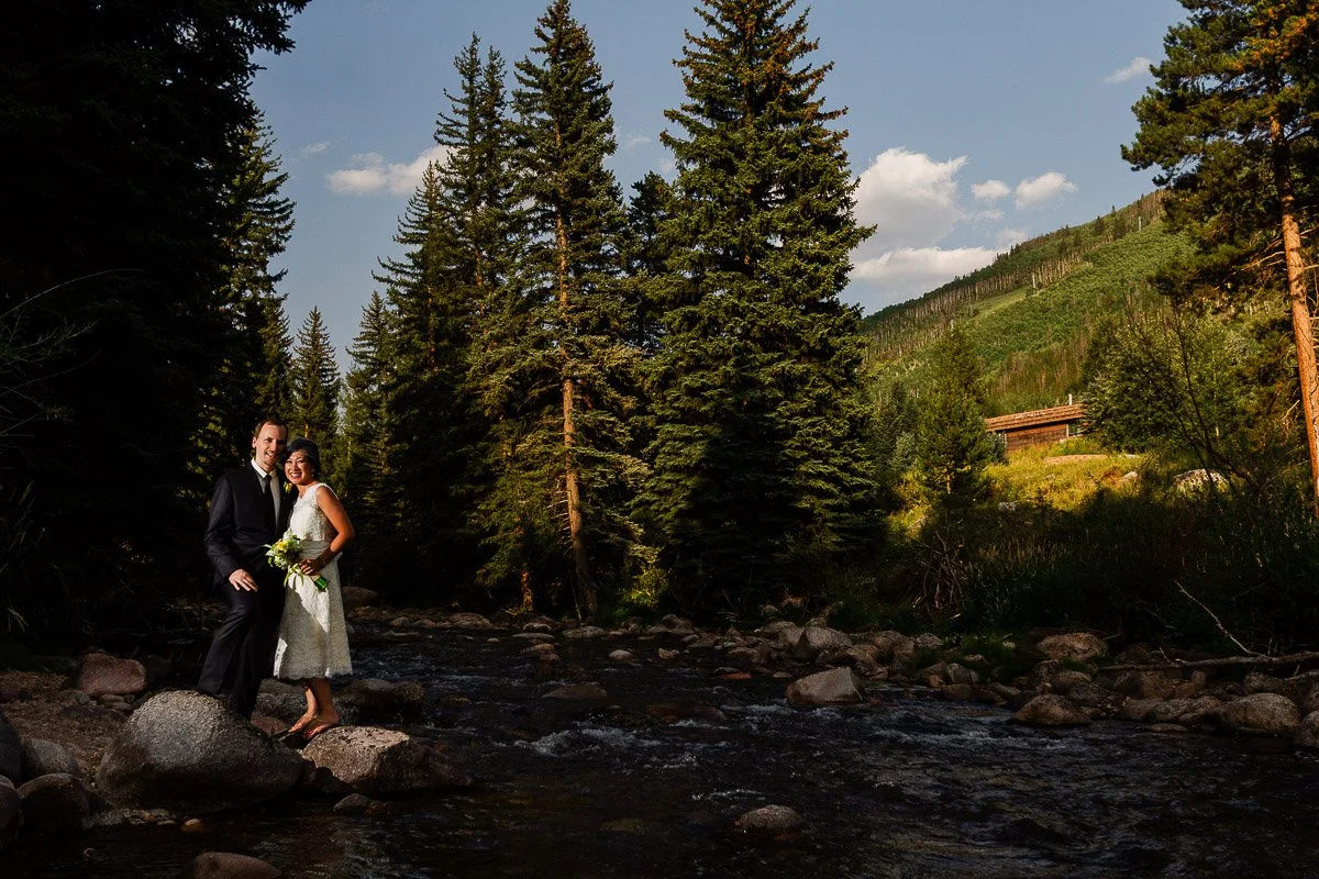 A couple in formal attire stands on rocks in Gore Creek, surrounded by tall evergreen trees under a clear blue sky. The scene conveys romance and tranquility during a Donovan Pavilion wedding in Vail, Colorado
