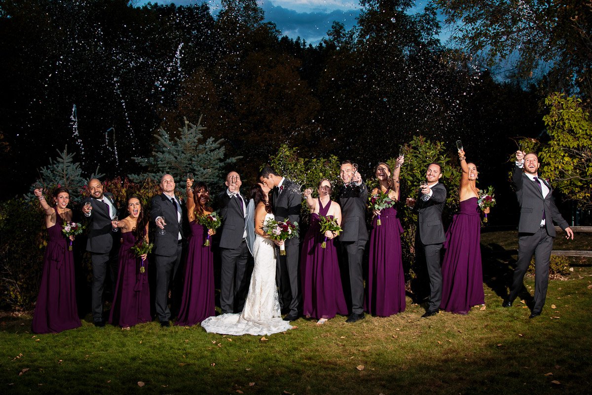 A joyous wedding party celebrates outdoors at night. The couple kisses in the center, surrounded by bridesmaids in purple dresses and groomsmen in suits, all spraying champagne.