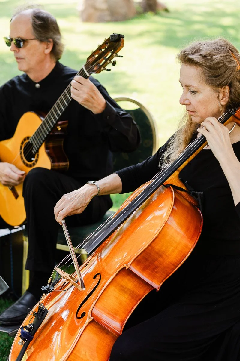 A woman intensely plays a cello, her bow gliding over the strings, beside a man in sunglasses playing a guitar. They're outdoors on green grass.