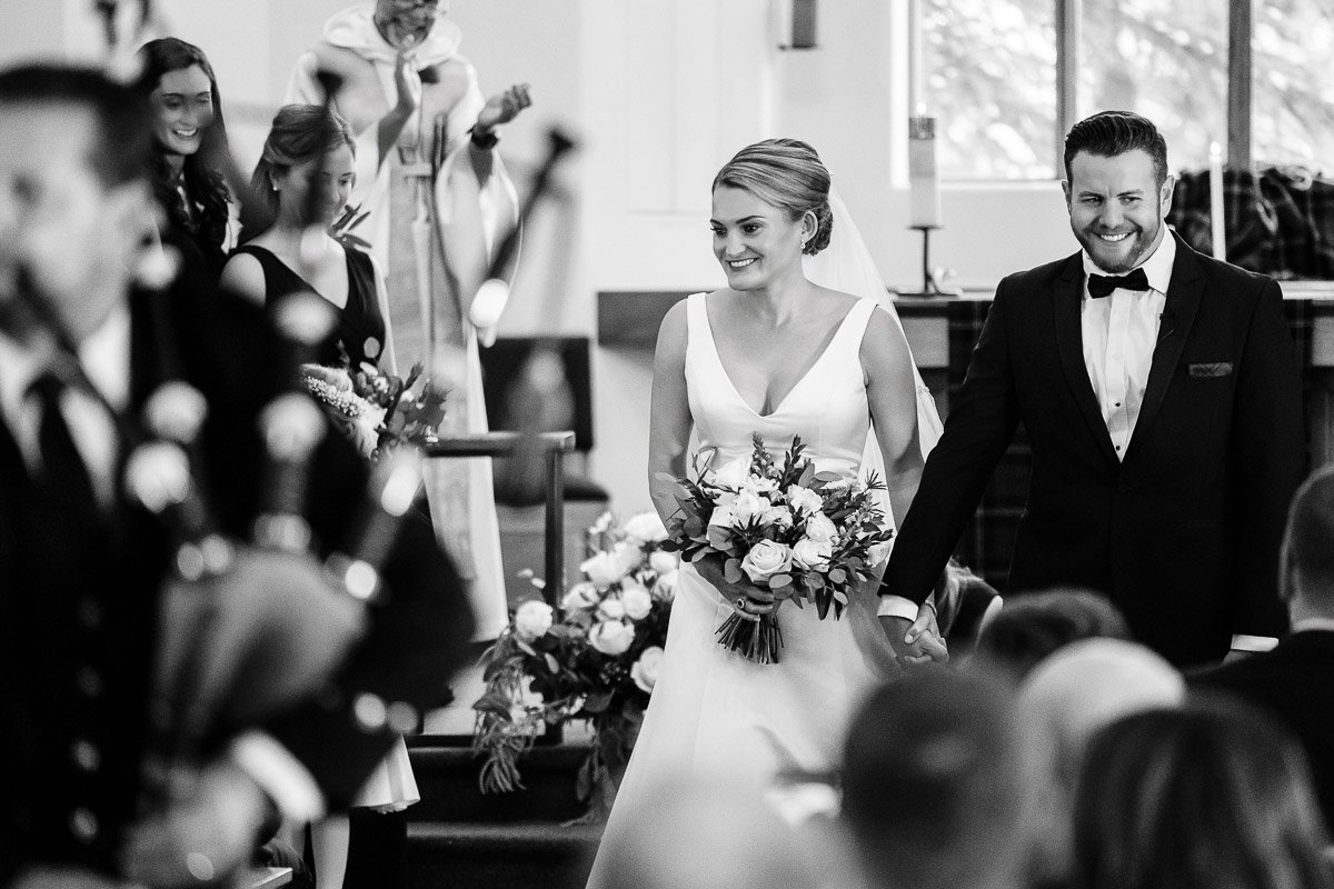 A joyful bride and groom walk down the aisle in a church, holding hands and smiling, while guests and a bagpiper are seen in the foreground.
