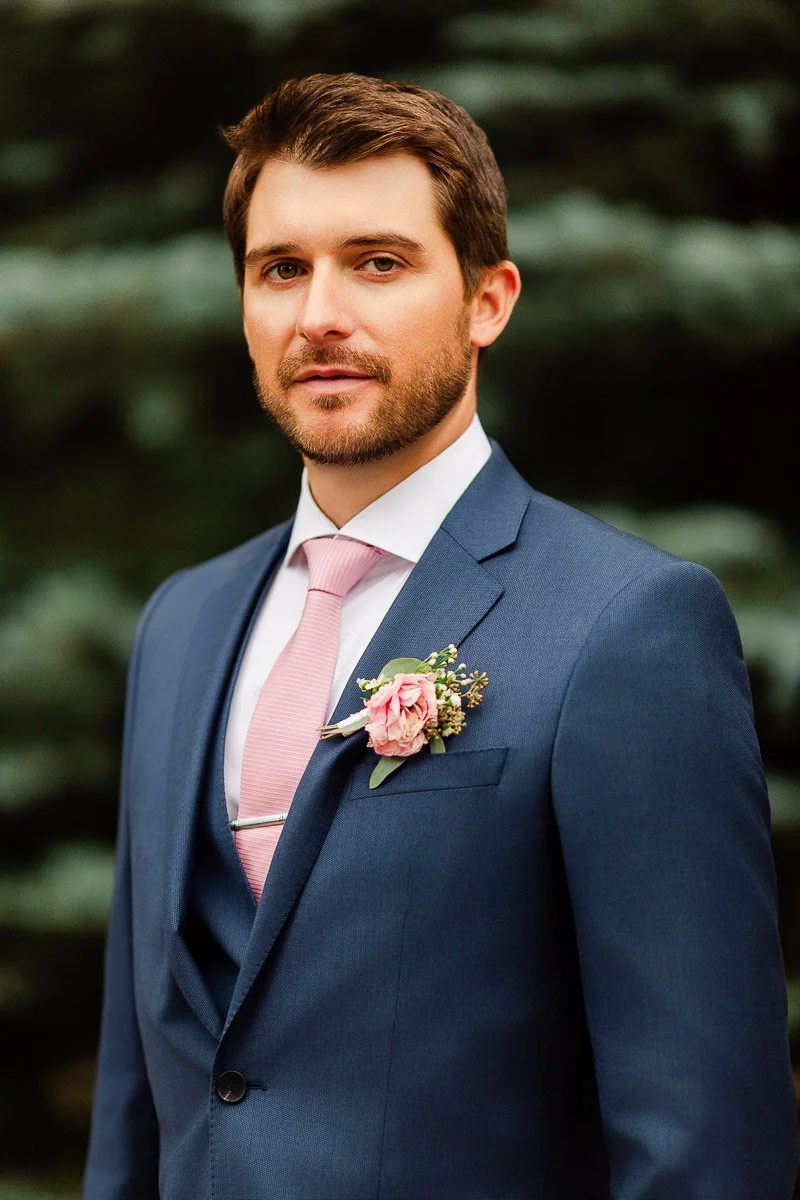 A man in a blue suit with a pink tie and floral boutonniere stands confidently, set against blurred green foliage, conveying elegance and calm.