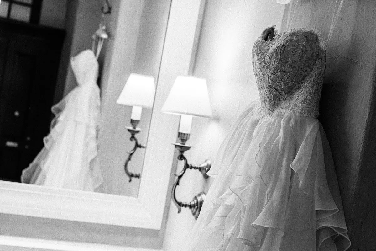 Black and white image of an elegant wedding dress with lace bodice and layered skirt hanging near a mirror. Soft lighting from wall lamps creates a serene, romantic mood.