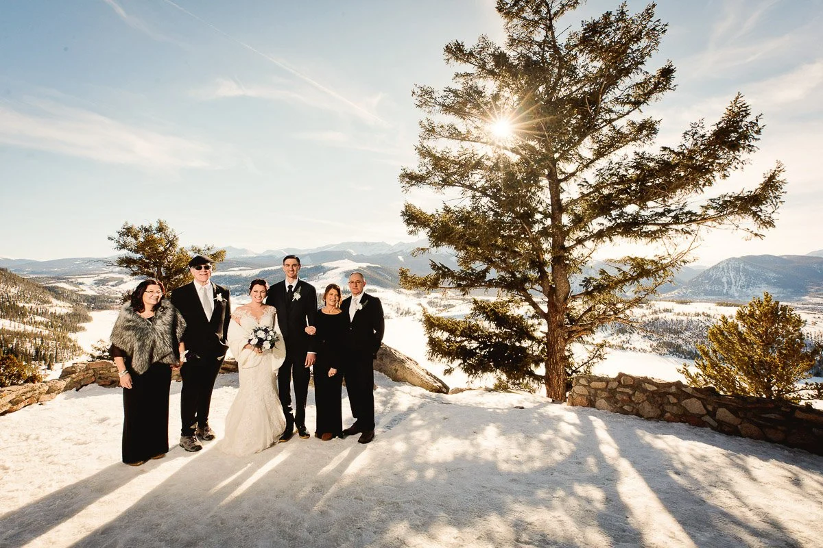A joyful wedding group stands on a snowy mountaintop, illuminated by a bright sun peeking through a tall tree. Scenic mountains form the backdrop.