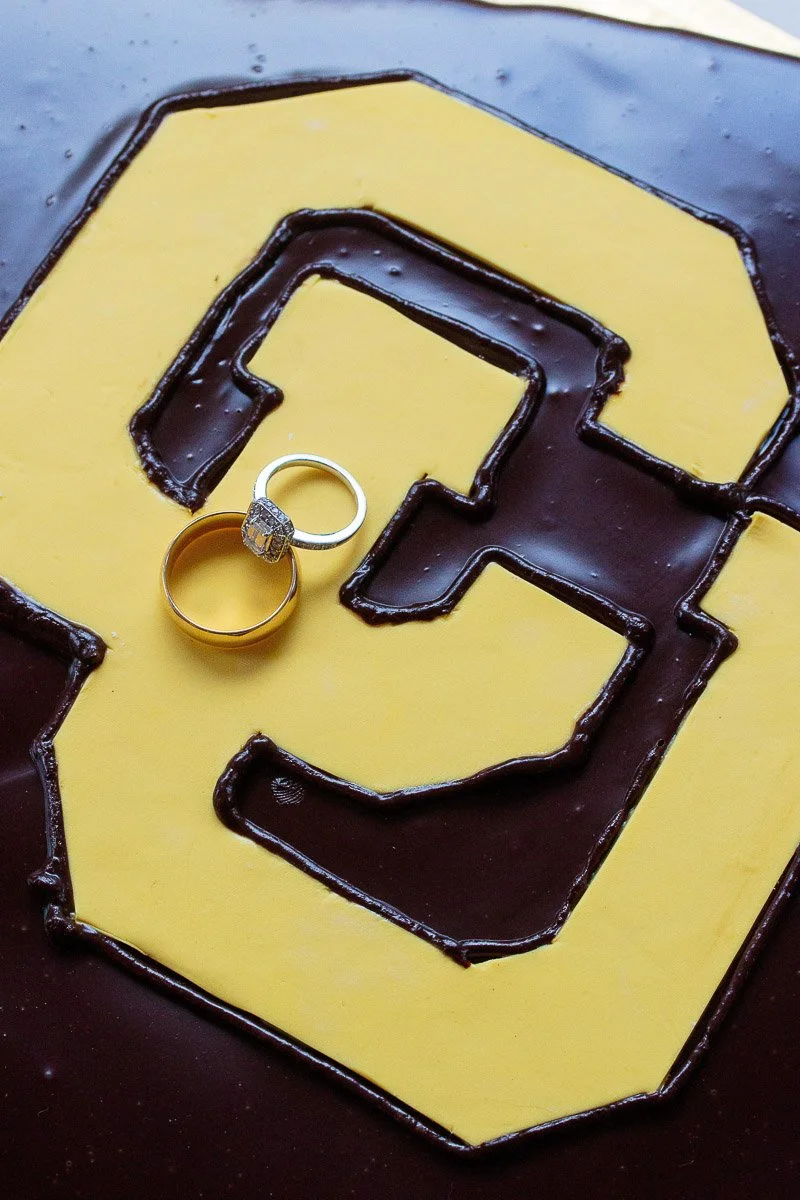 Two rings, one gold band and one with a diamond, rest on a chocolate cake decorated with a large Colorado University Buffs logo outlined in dark chocolate.