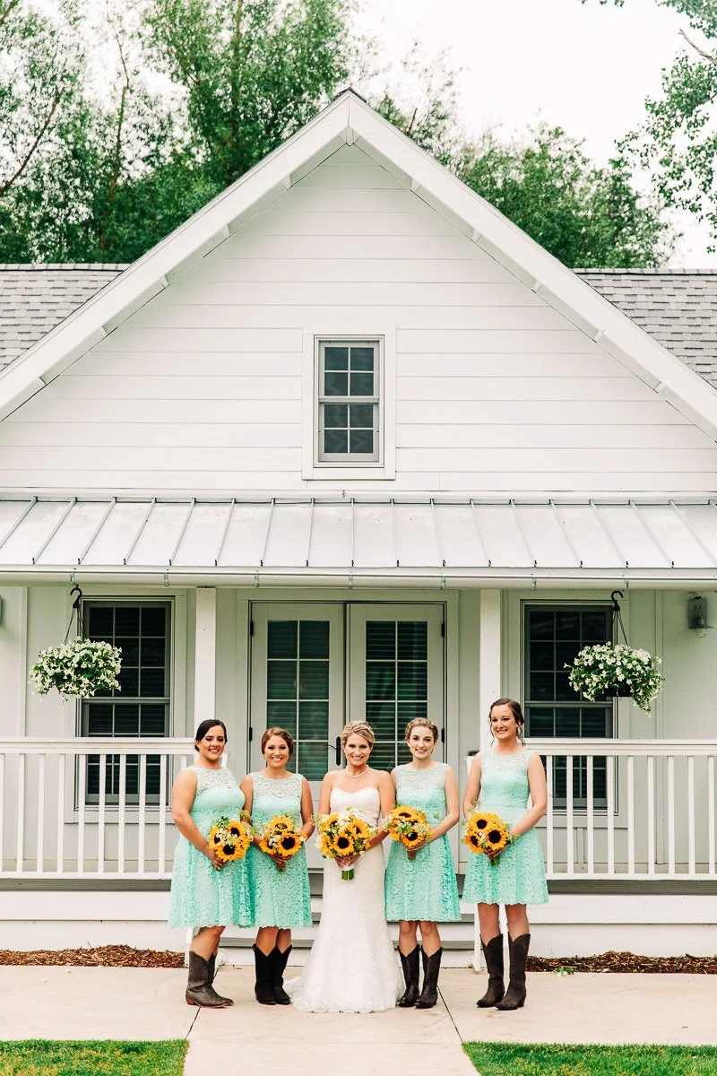 Bride in white gown and four bridesmaids in teal dresses and brown boots, holding sunflower bouquets, stand before a white house with green plants during a Barn at Raccoon Creek wedding in Littleton, Colorado