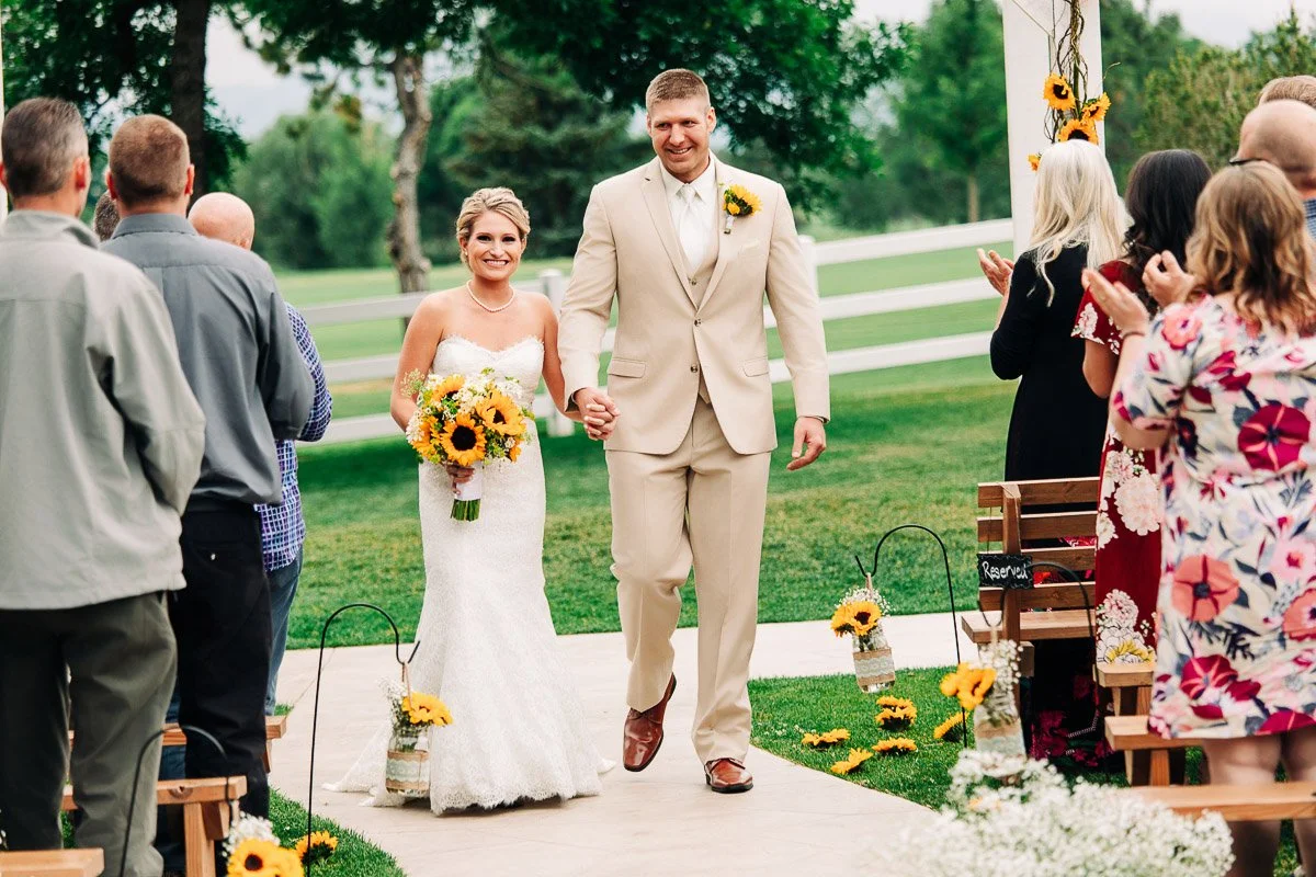 Bride and groom cheerfully walk down an outdoor aisle adorned with sunflowers. Guests on either side applaud. The setting is lush and green.