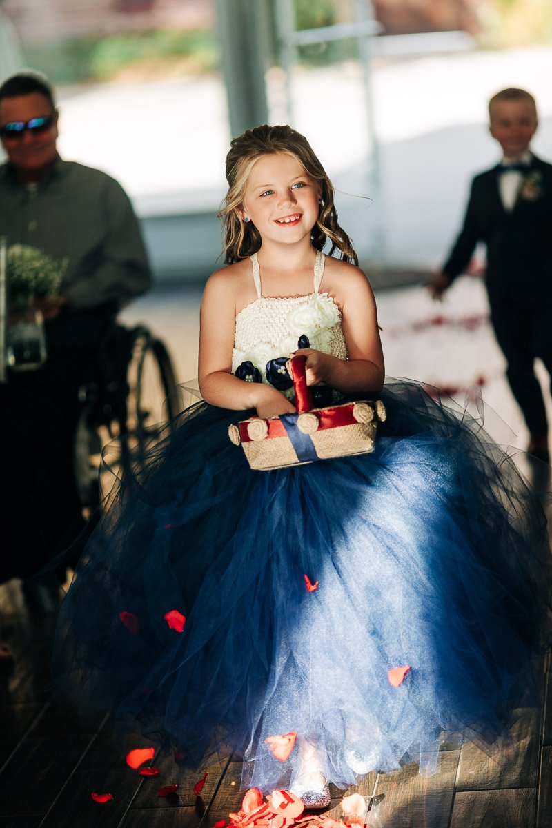 Young girl in a blue tulle dress joyfully scatters red rose petals from a basket, accompanied by wedding guests, including a boy in a suit. Bright, celebratory atmosphere.