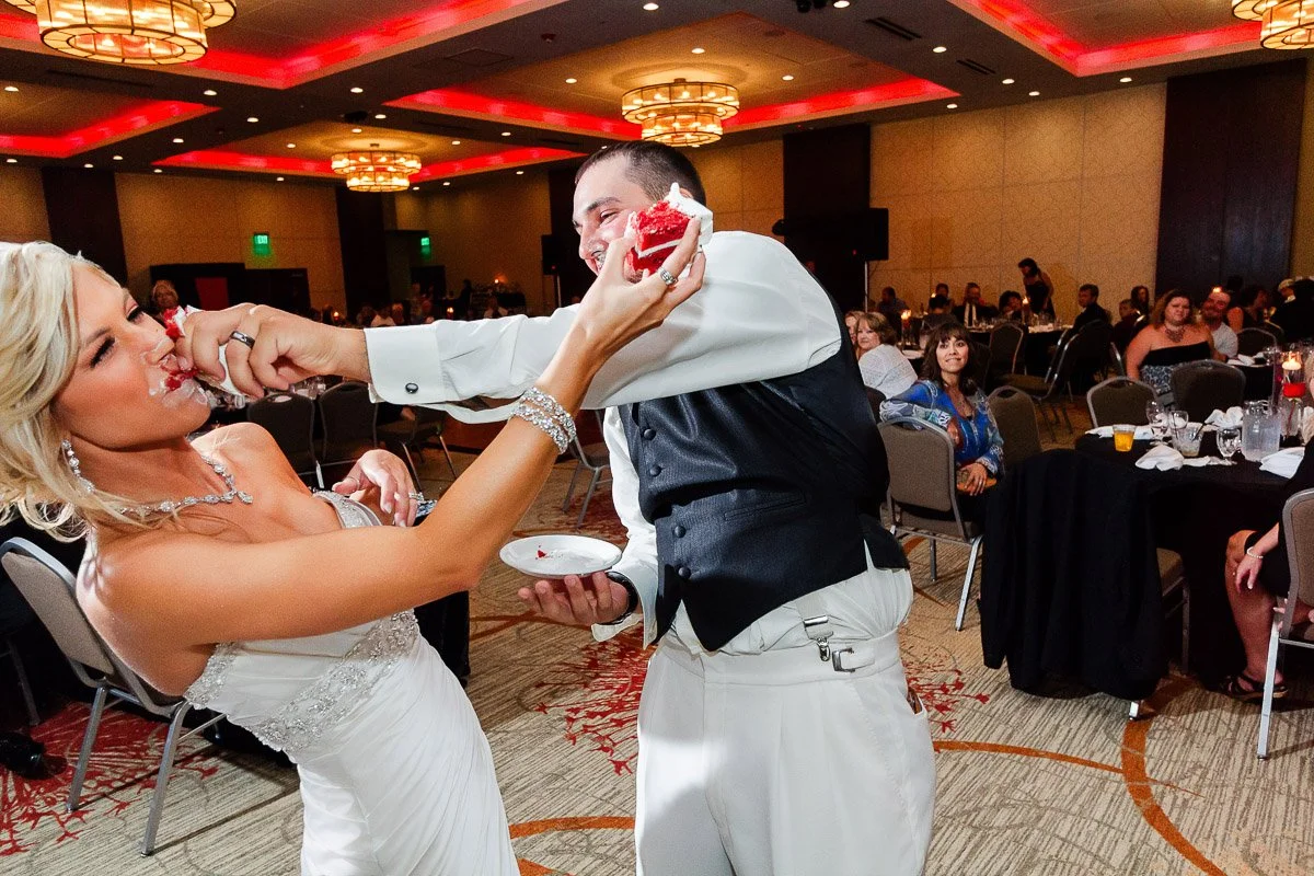 A bride playfully smears cake on the groom's face in a lively wedding reception. Guests watch with amusement. The room is warmly lit with elegant chandeliers.