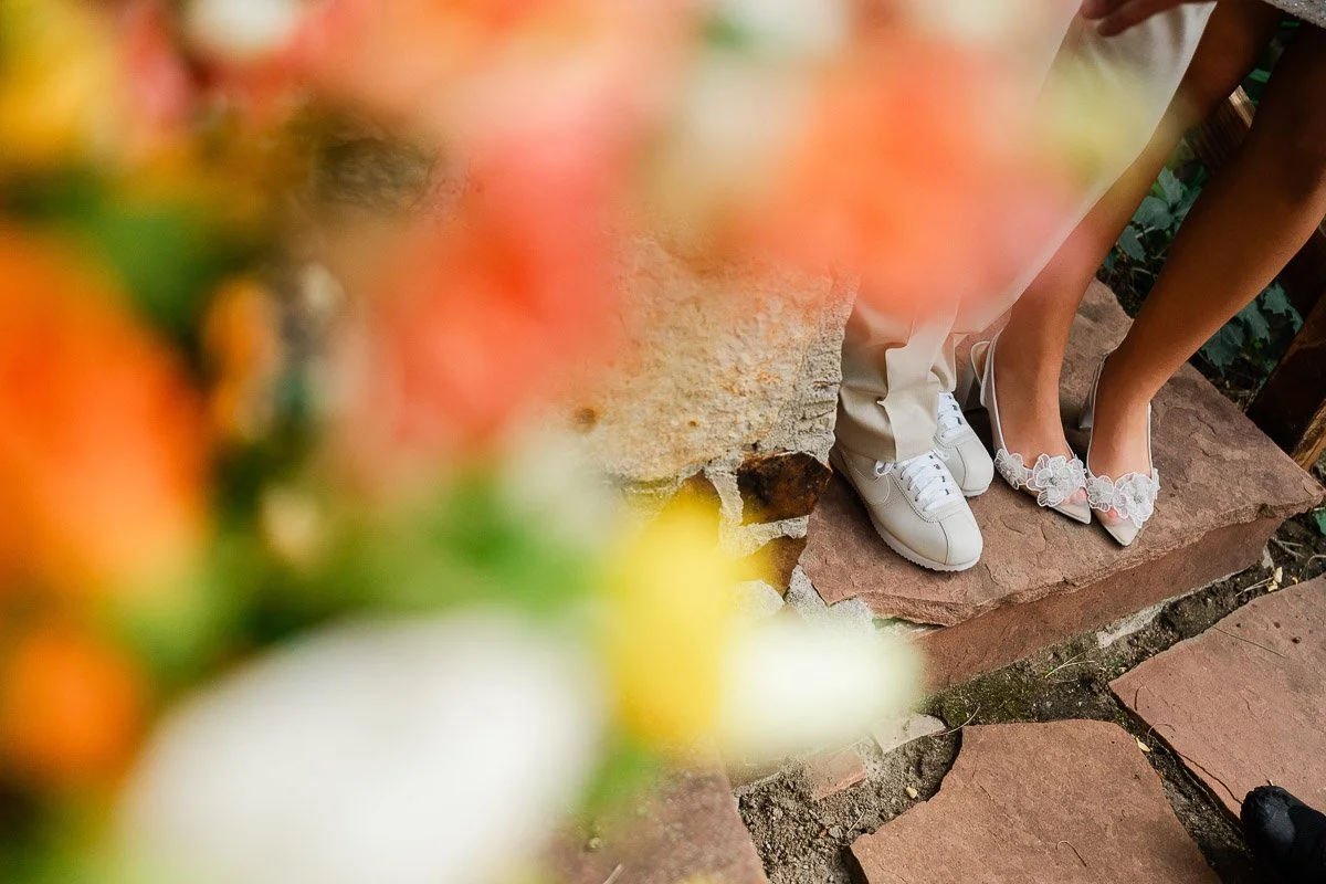 Close-up of a couple's feet, one in white sneakers and the other in floral heels, standing on stone steps. Blurred colorful flowers in the foreground.