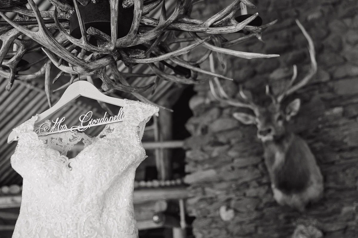 A lace wedding dress hangs on a wooden hanger labeled "Mrs." under an antler chandelier, with a mounted deer head on a rustic stone wall in the background.