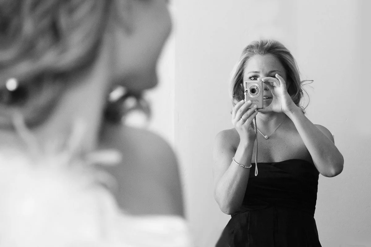A woman in a strapless dress smiles and holds a camera up to her eye. Her reflection shows another smiling woman. The scene conveys joy and connection.