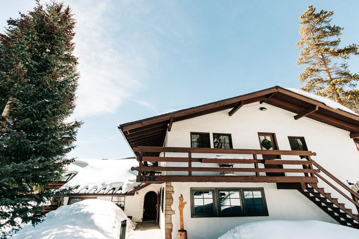 A cozy chalet-style house with a snow-covered roof and wooden balcony sits under a clear blue sky. Tall evergreen trees frame the peaceful winter scene at Ski Tip Lodge in Keystone Colorado