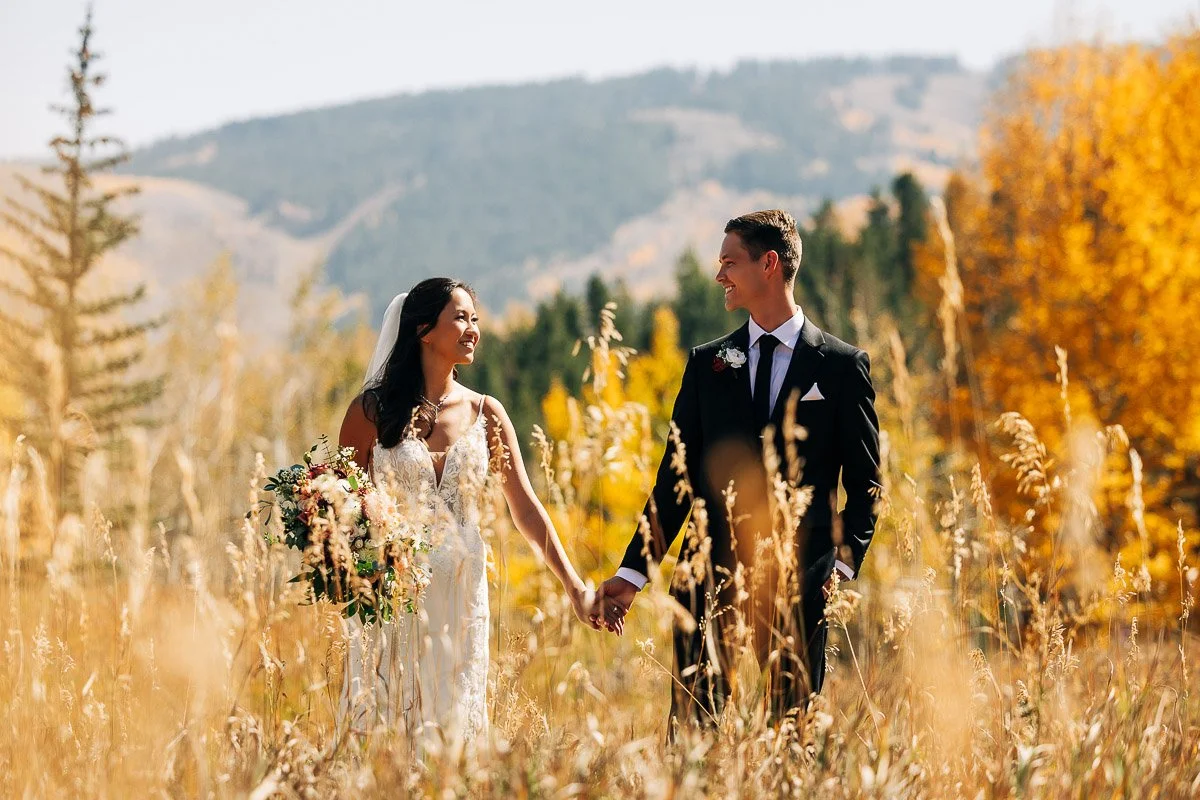 Bride and groom hold hands, smiling at each other in an autumnal meadow. The scene is framed with golden grasses and colorful fall foliage.