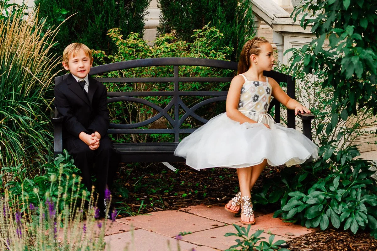 A young boy in a black suit and a girl in a white dress sit on a decorative bench in a lush garden. The mood is formal yet playful.