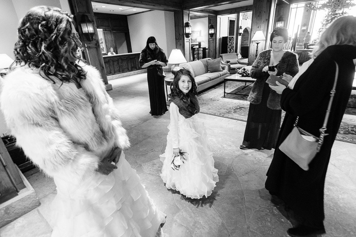 A young girl in a white dress smiling amid adults in formal wear, inside an elegant, warmly lit hotel lobby, creating a joyful atmosphere.