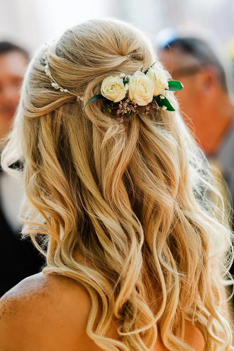 Blonde hair styled in soft waves with a half-updo adorned by white roses and greenery. The scene conveys elegance and a joyful, celebratory atmosphere.