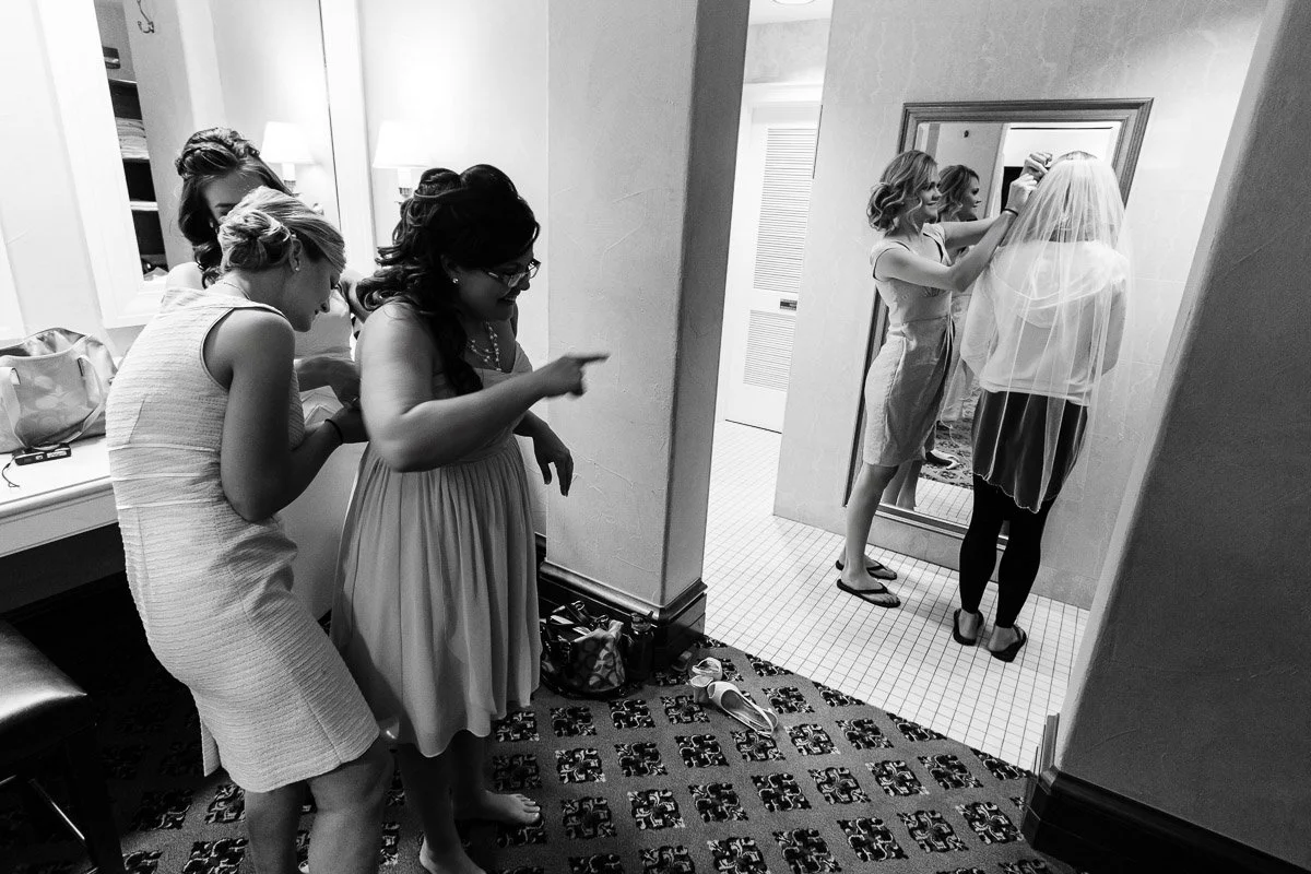 A group of women in a dressing room, some smiling and pointing as one adjusts a bride's veil in front of a mirror. The scene is joyful and bustling.
