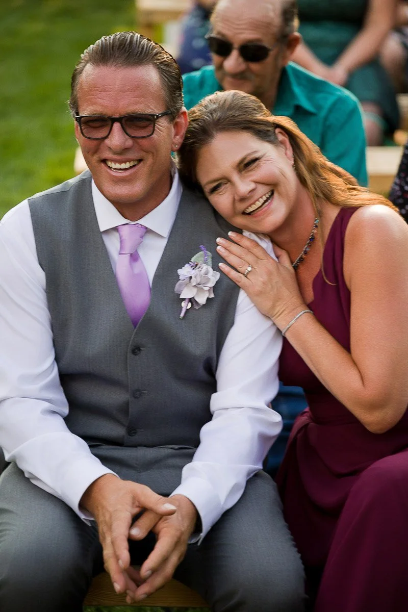 A smiling couple seated outdoors, the man in a gray suit and lavender tie, and the woman in a burgundy dress. She rests her head on his shoulder.