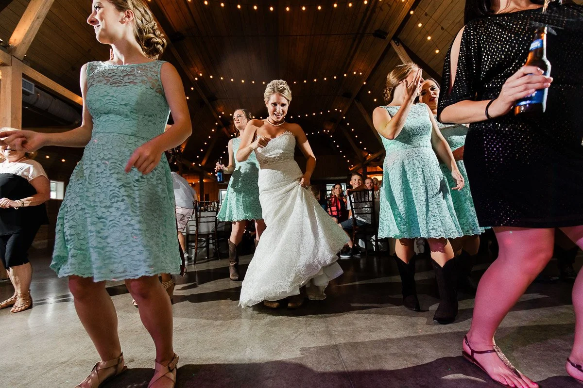 A joyful bride in a white gown dances energetically with bridesmaids in mint dresses at a rustic wedding reception under string lights.