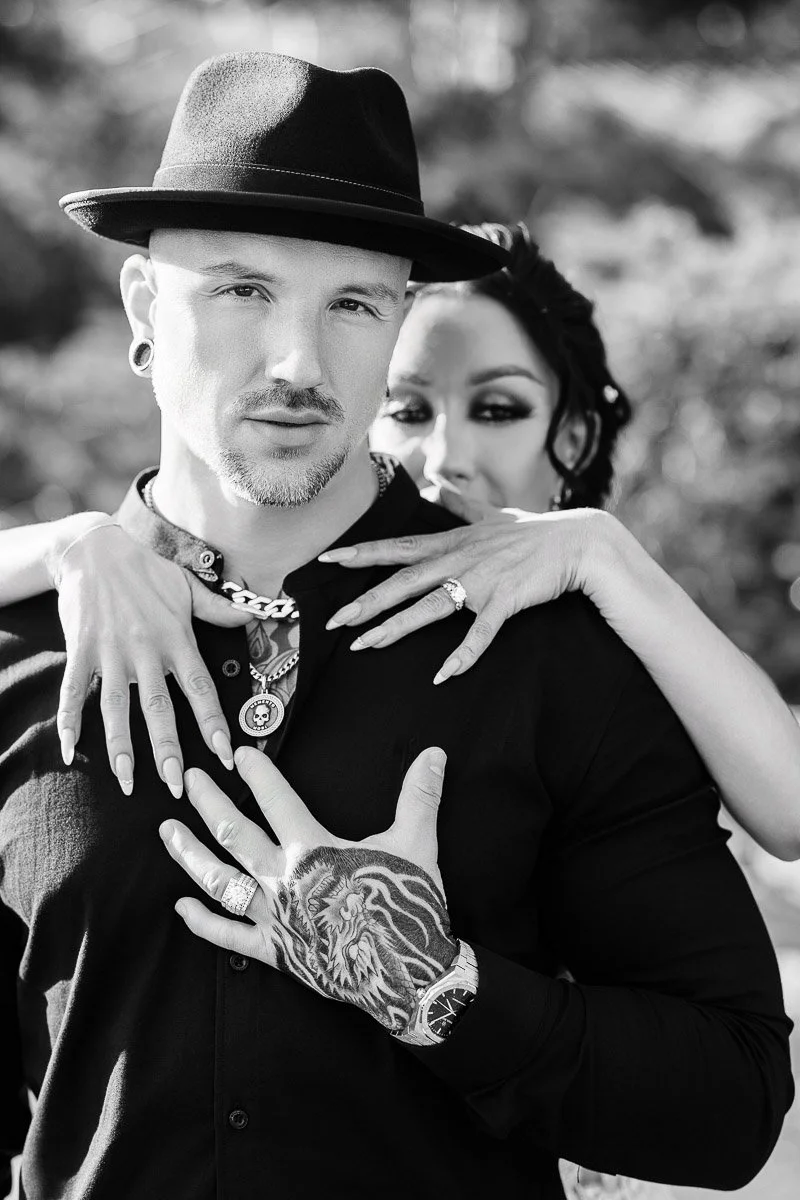 Black and white photo of a confident man in a hat and patterned tattoo, embraced from behind by a woman, evoking elegance and connection.