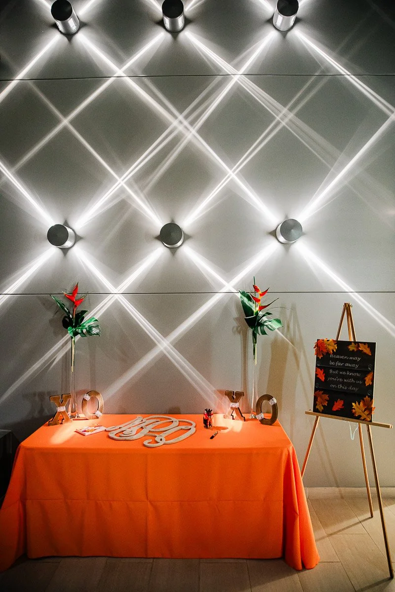 Orange table with "XO" letters and tropical flowers, under geometric wall lights. A fall-themed sign on an easel conveys warmth and celebration.