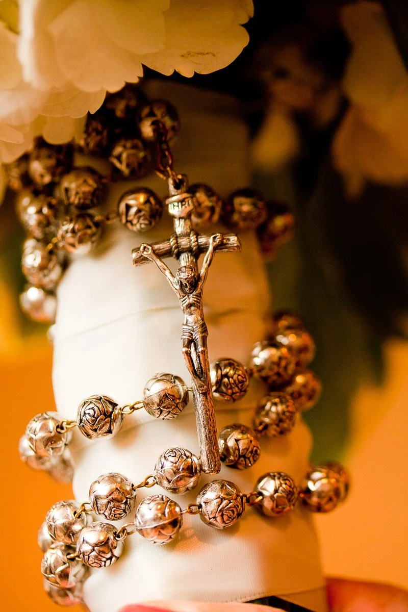 Close-up of a silver rosary with rose-shaped beads and crucifix, wrapped around white fabric. Soft, warm lighting creates a serene, contemplative mood.