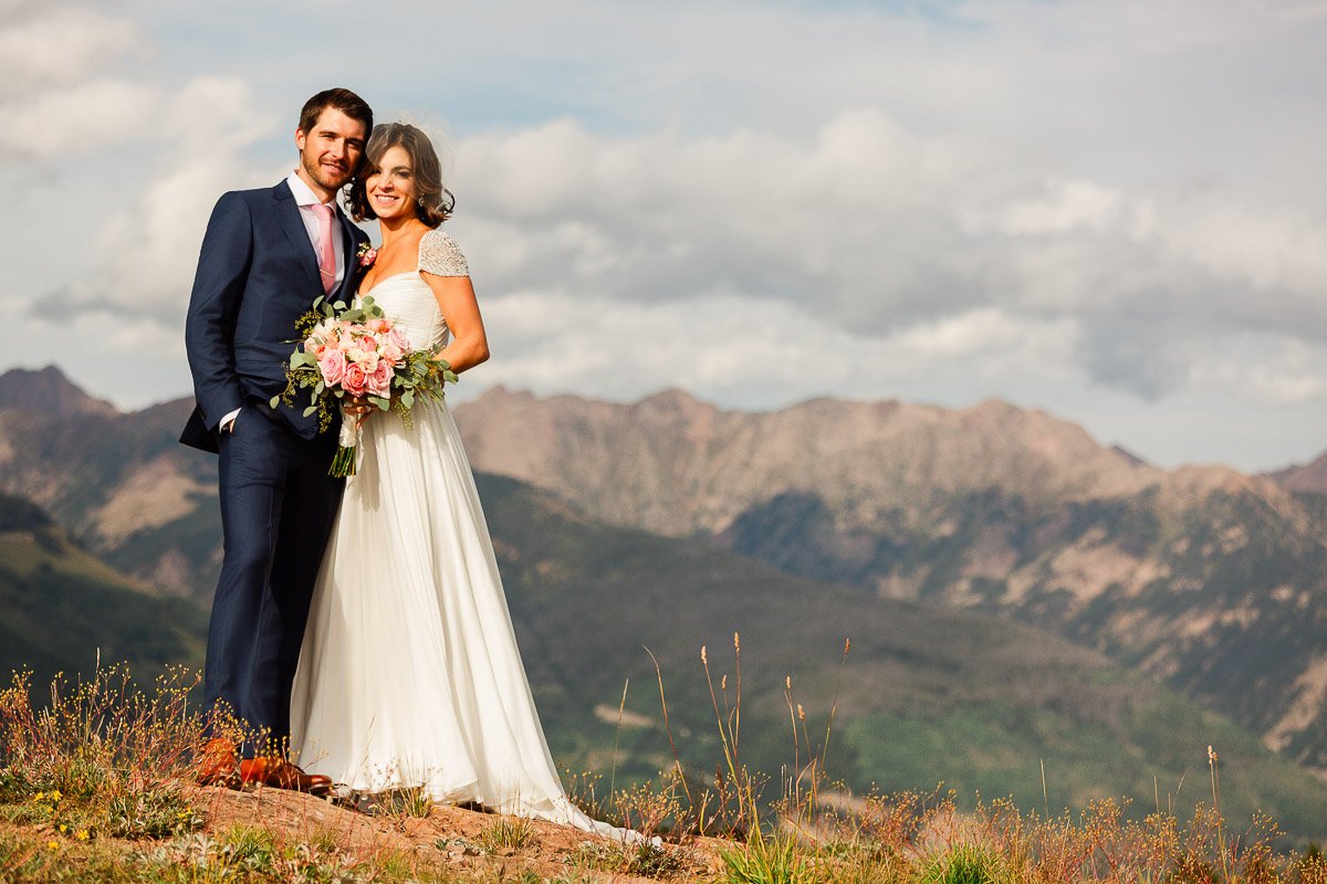 A bride and groom smile joyfully on a mountain, with rugged peaks of the Gore Mountain Rangeand a cloudy sky in the background. She holds a bouquet of pink flowers during a Four Seasons Resort wedding in Vail Colorado