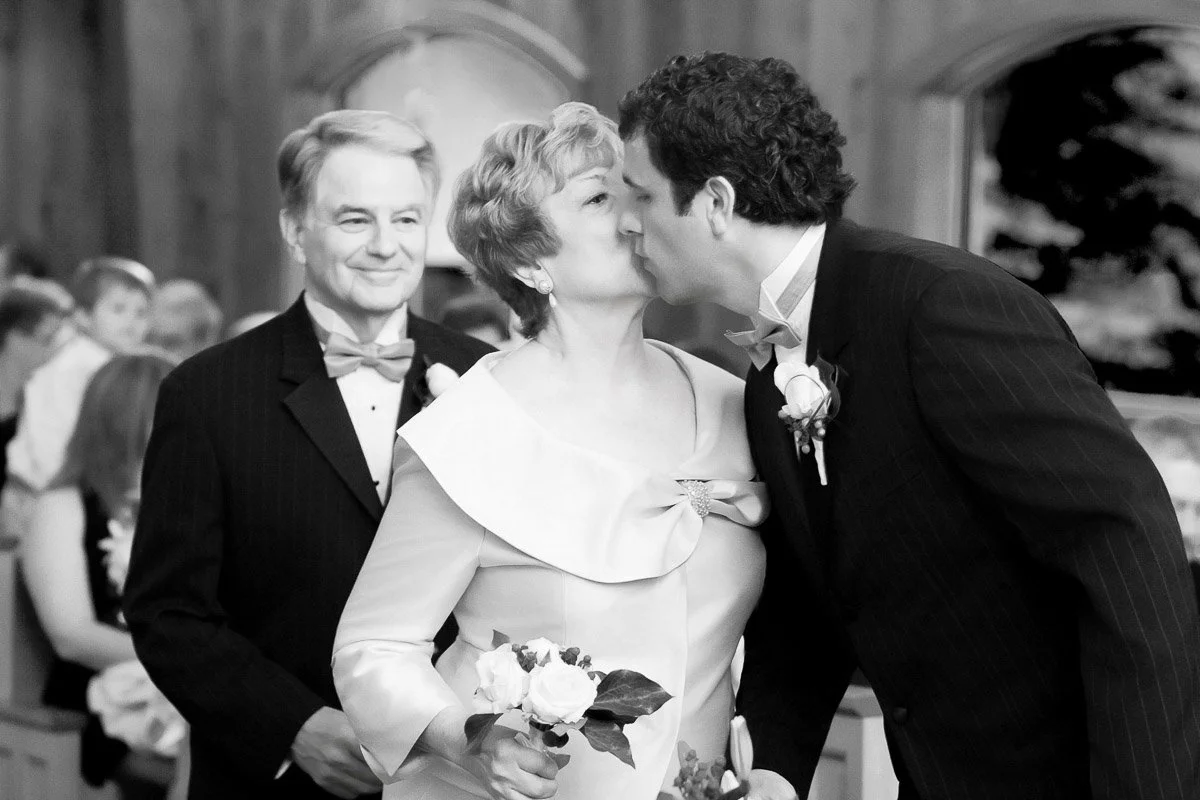 A joyful wedding scene in black and white. A woman holding flowers receives a kiss on the cheek from a man in a suit, while another man smiles beside them.