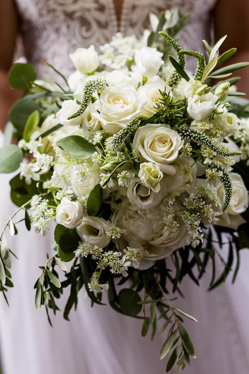 A bridal bouquet features a lush arrangement of white roses, delicate blossoms, and greenery, conveying elegance and a romantic, serene tone.