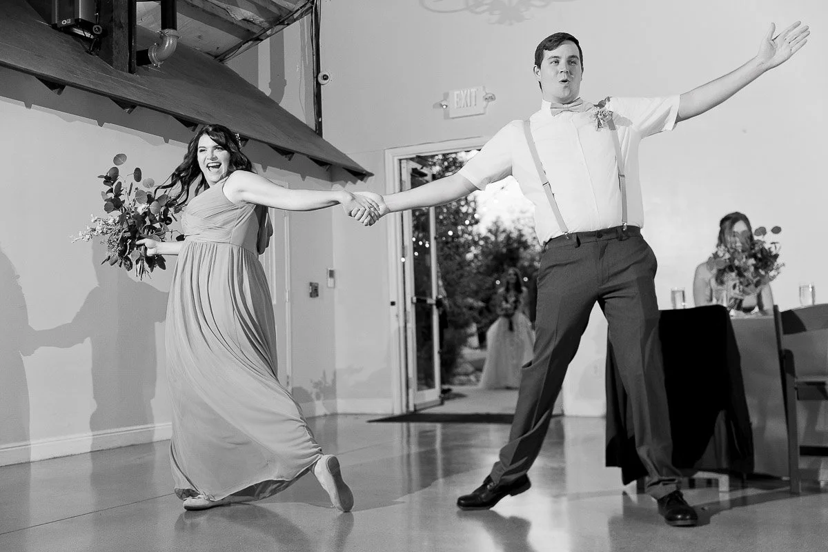 A bridesmaid and groomsman joyfully dance hand-in-hand indoors. She's in a flowing dress, holding flowers, both smiling, conveying a festive mood.