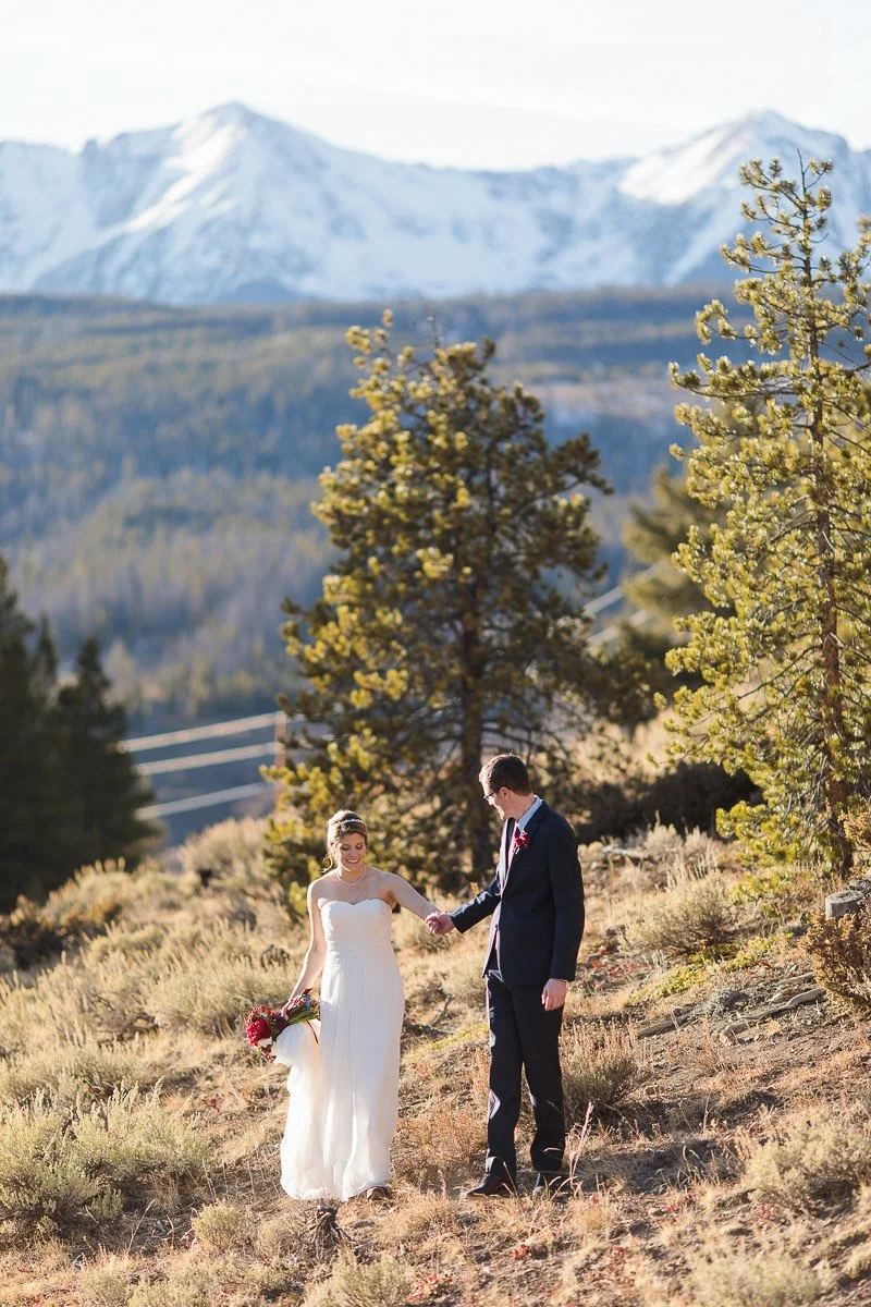 Bride and groom walk hand in hand on a mountainside with snow-capped peaks in the background. She holds red flowers, creating a joyful, serene scene during a The Lodge at Breckenridge wedding.
