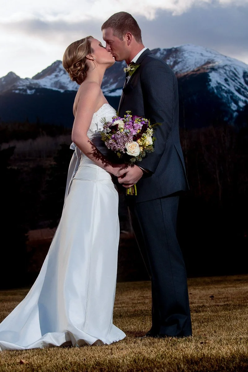 Bride in white gown and groom in dark suit kiss with mountains in the background. The bride holds a bouquet. The scene is romantic and serene.
