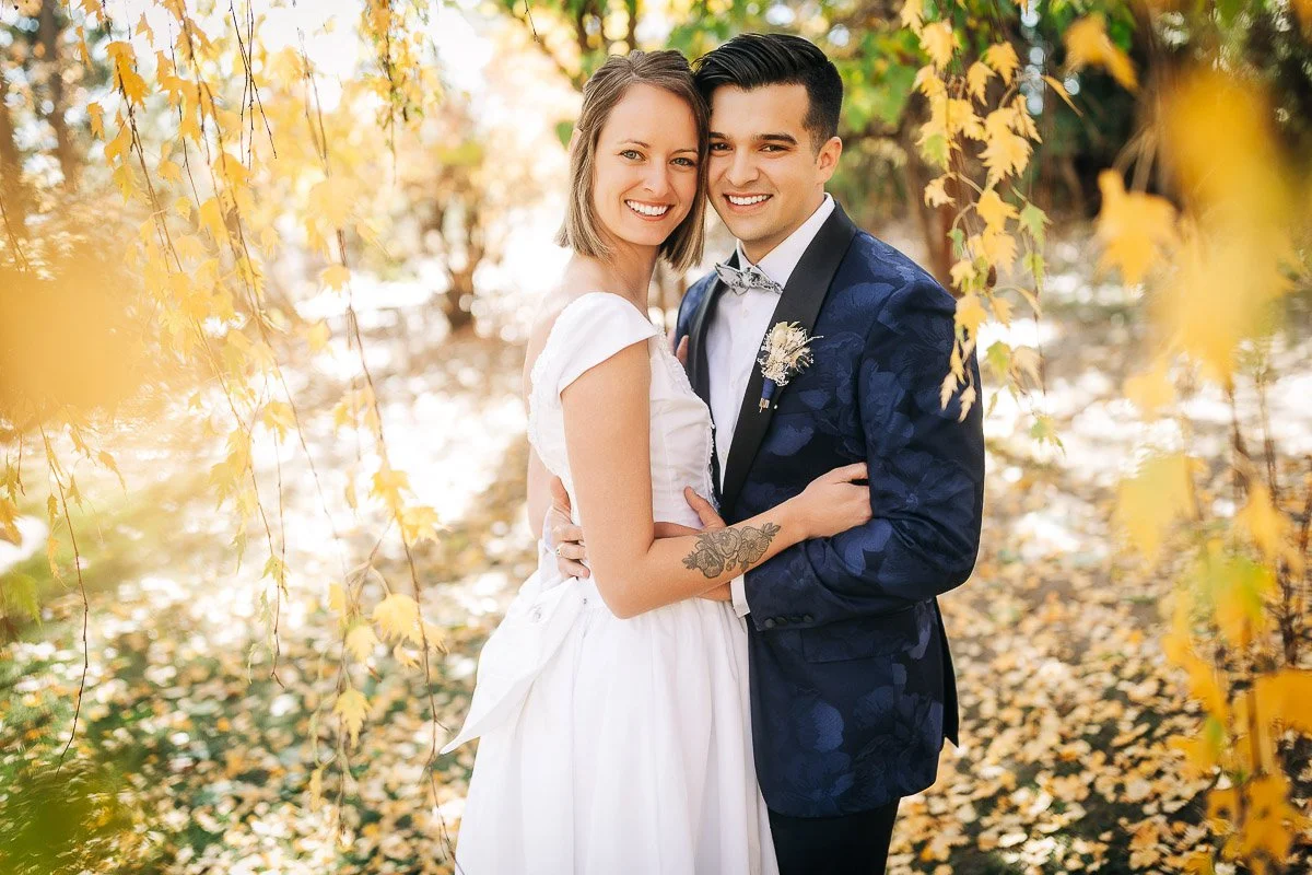 A joyful couple embraces under a canopy of golden autumn leaves. The woman wears a white dress, the man a floral-patterned suit, both smiling warmly.