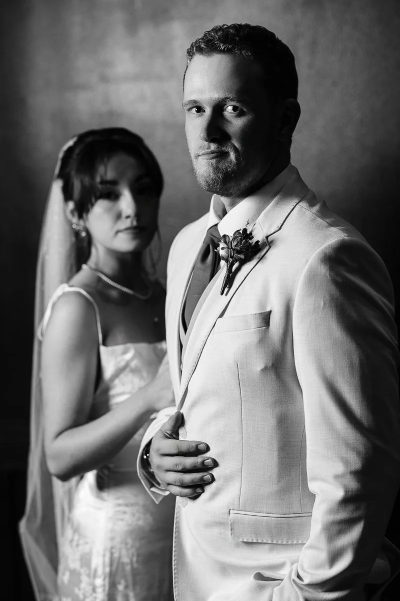 A bride and groom in formal attire stand together, exuding elegance and romance. The groom wears a light suit, the bride a veil, both gazing softly. Black and white.